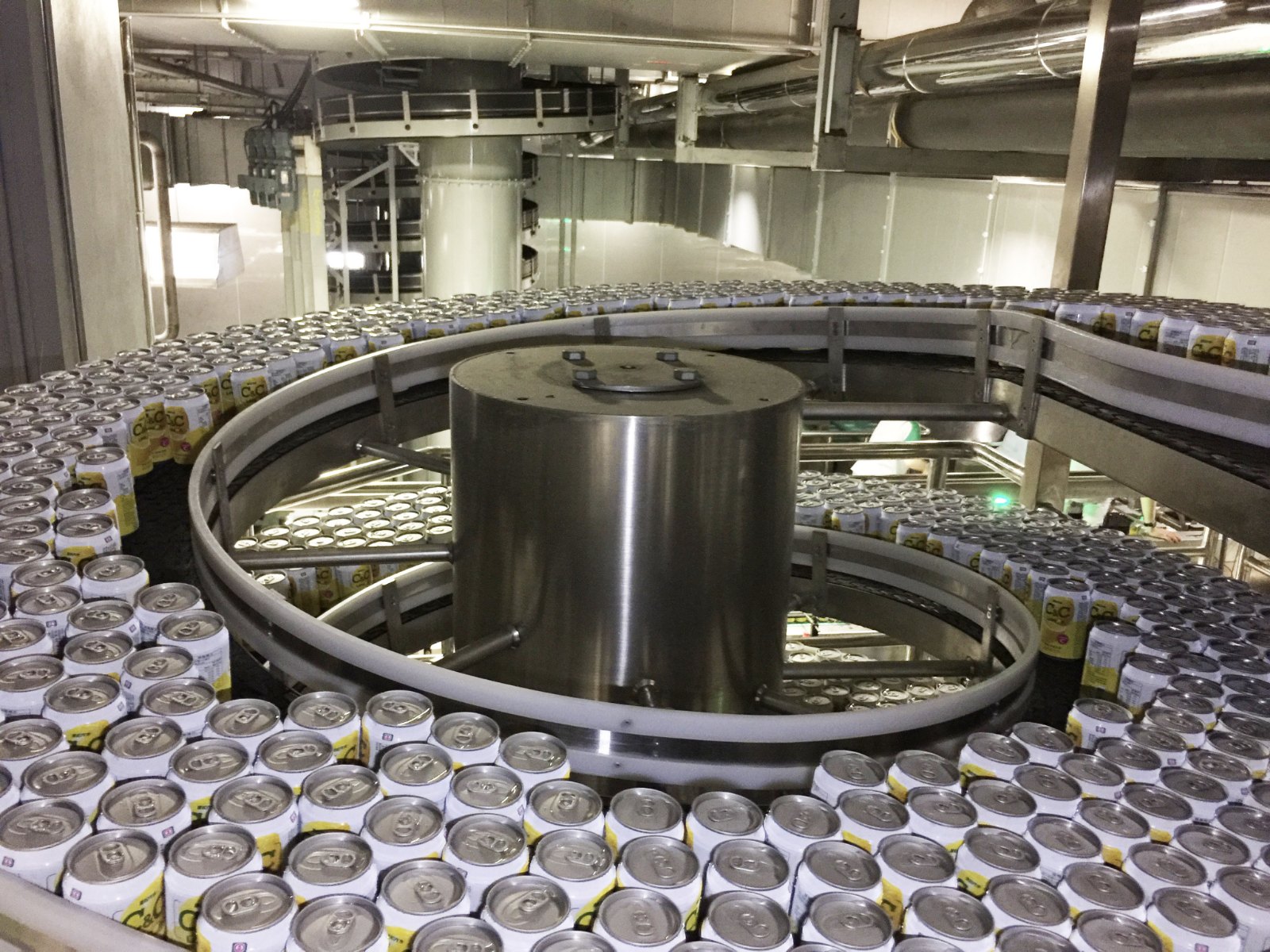Beverage cans standing in a dense queue across the full width of a spiral accumulation conveyor track, wound around a stainless steel column.
